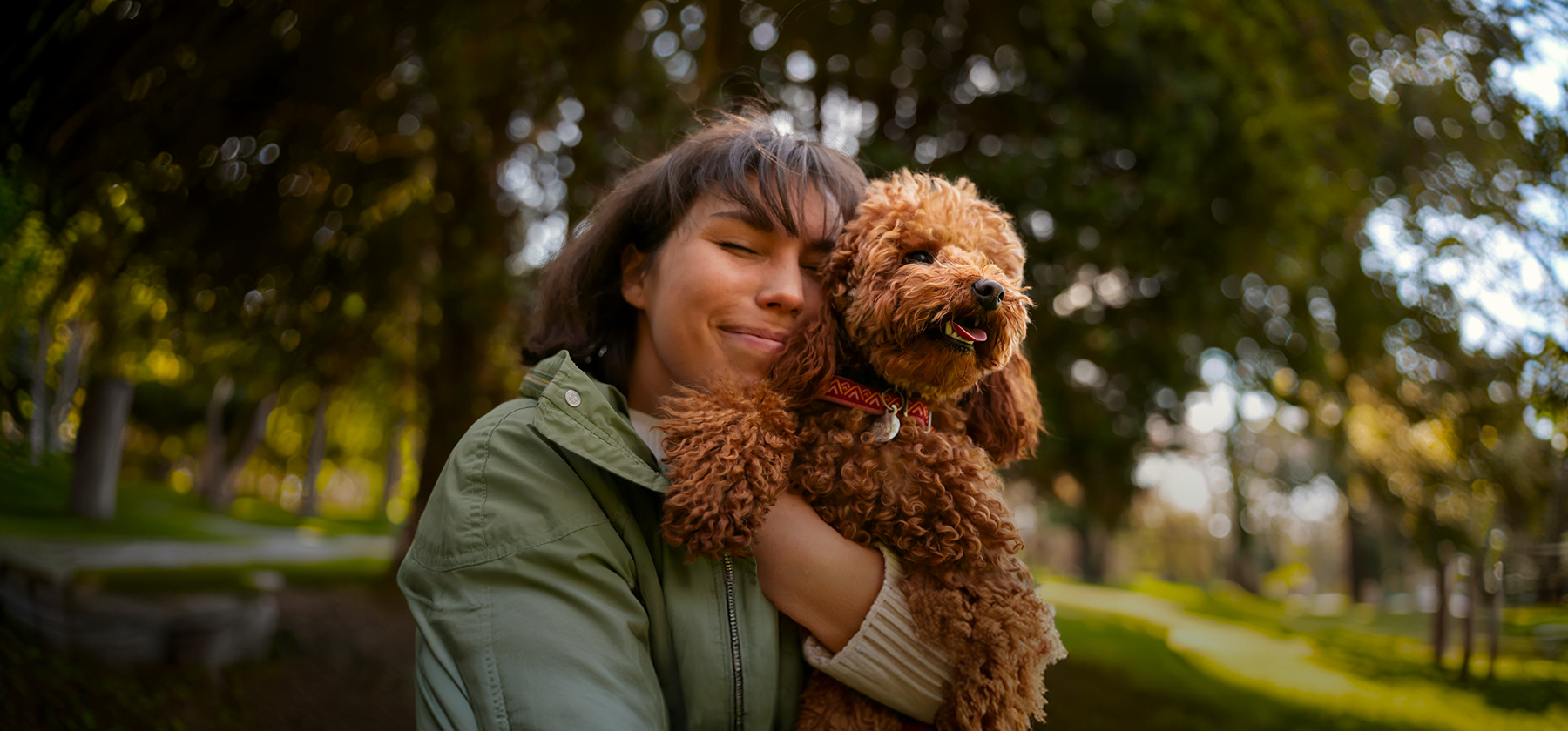 A woman in a green jacket joyfully cuddling a fluffy golden-brown poodle in a sunny park in Germany.
