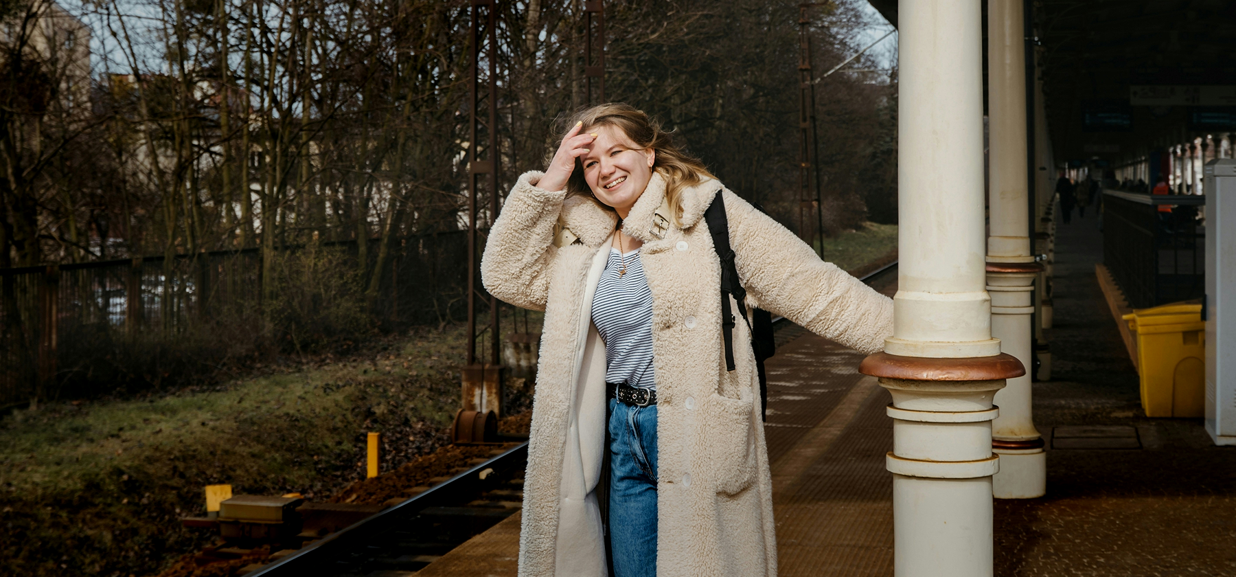 A young woman in a fluffy cream coat smiling cheerfully on a train platform with a backpack, her hair blowing in the wind in Germany.