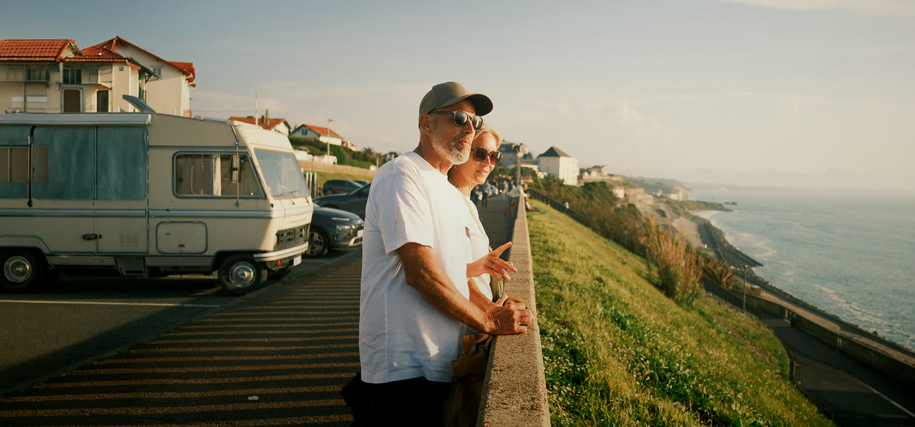An older couple wearing sunglasses leaning on a wall overlooking a coastal road and the sea at sunset in France.
