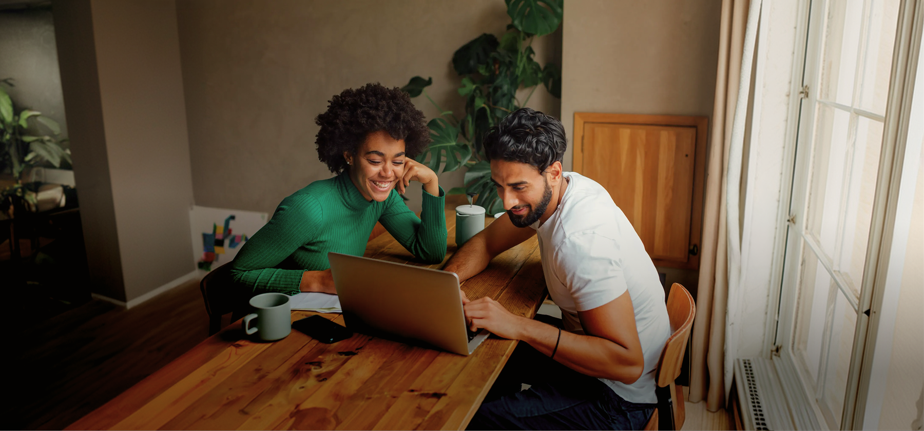 A smiling couple looking at a laptop together at a wooden dining table with coffee cups in a plant-filled apartment in Spain.