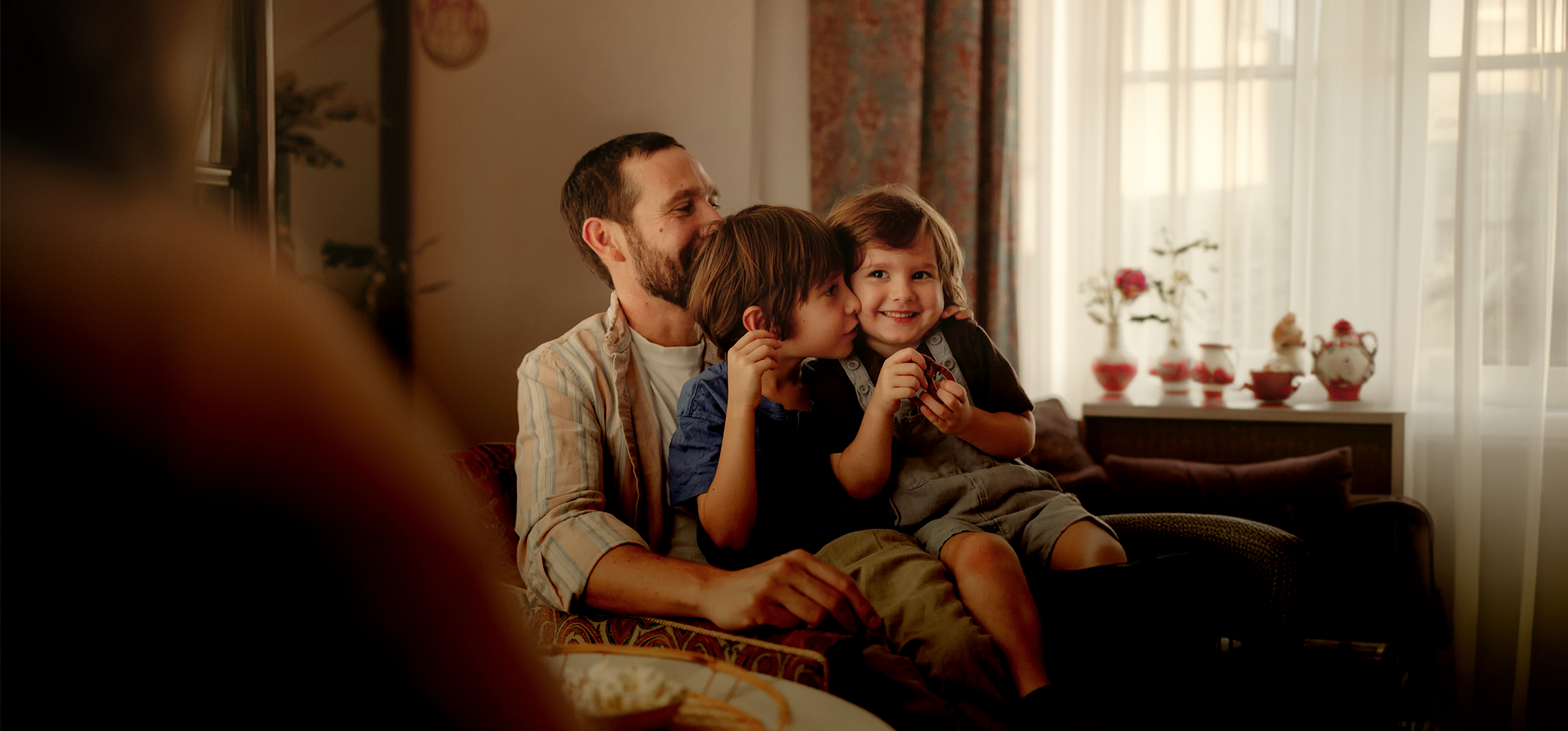 A father sitting on a sofa with two young children cuddling up to him in a warmly lit living room in Germany.
