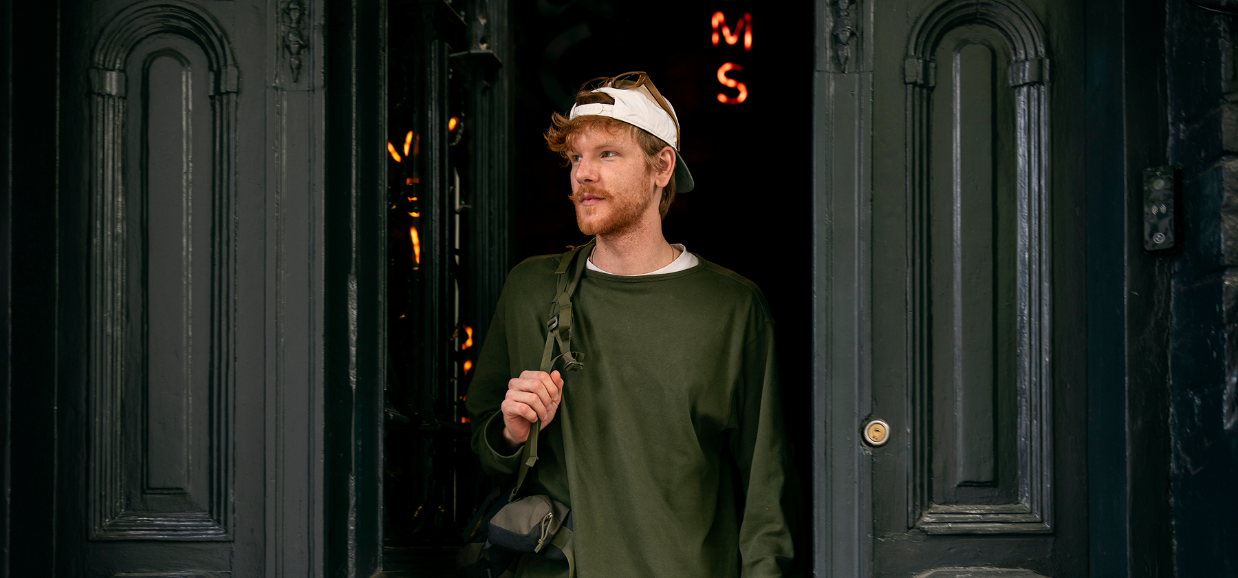 A man wearing a cap and carrying a green backpack walks out through a doorway onto a city street.