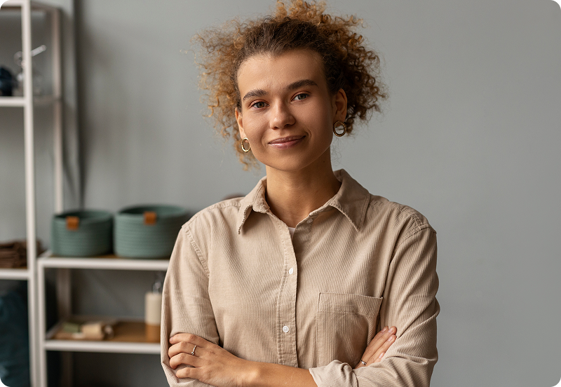 Image of a woman with curly hair, smiling confidently with arms crossed, wearing a beige shirt in a minimalist room.