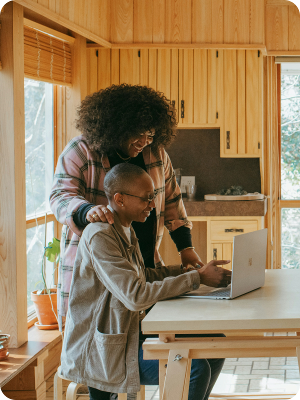 Two women smiling and looking at a laptop together at a table in a warm wooden-panelled kitchen, one seated and one standing with her hand on the other's shoulder.