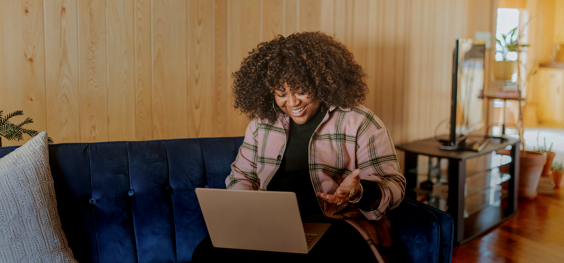 A woman with curly hair wearing a plaid jacket sitting on a blue velvet sofa, smiling and gesturing while on a video call on her laptop in a warm wooden-panelled living room.