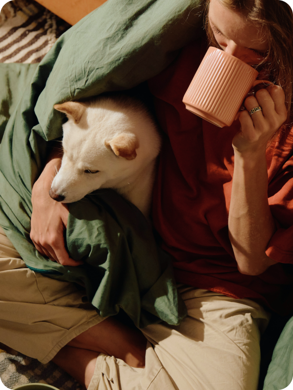 A person relaxing under a green blanket, holding a pink mug and cuddling a white dog in a cosy sunlit setting in Germany.