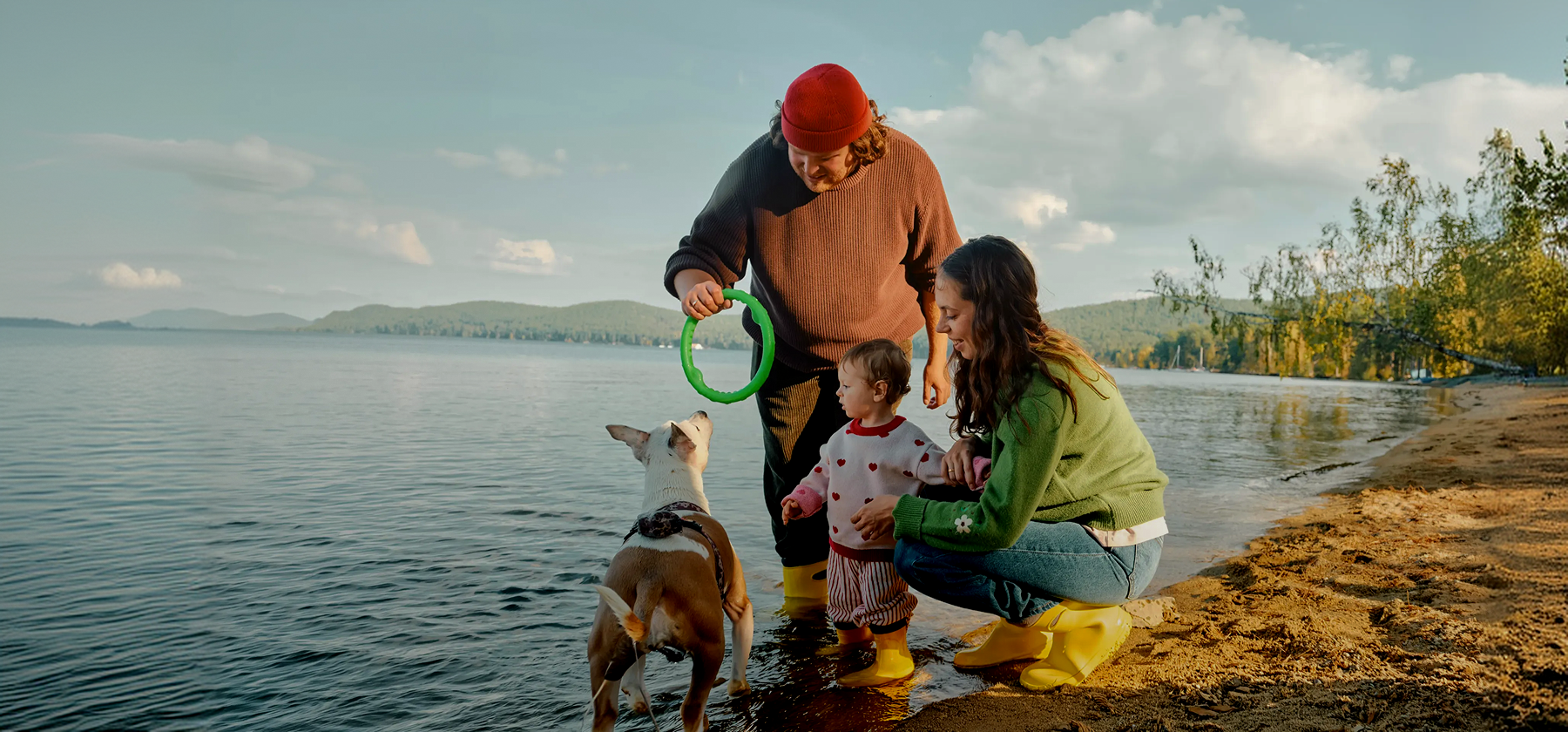 A young family with a toddler and a dog playing together on a lakeside shore, with a man holding a ring toy while a woman crouches beside the child in Germany.