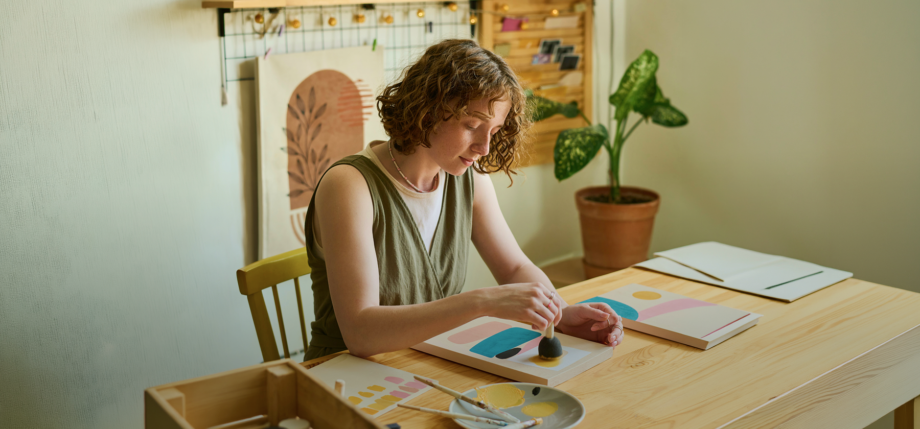 A young woman with curly hair working on colourful abstract artwork at a wooden desk in a bright, plant-filled creative studio in Germany.