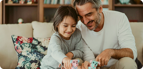 A father and young daughter sitting together on a sofa, smiling and playing with a toy in a cosy living room in Germany.