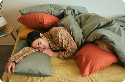A person lying in bed surrounded by colourful pillows in sage green, terracotta and mustard tones, resting peacefully in Germany.