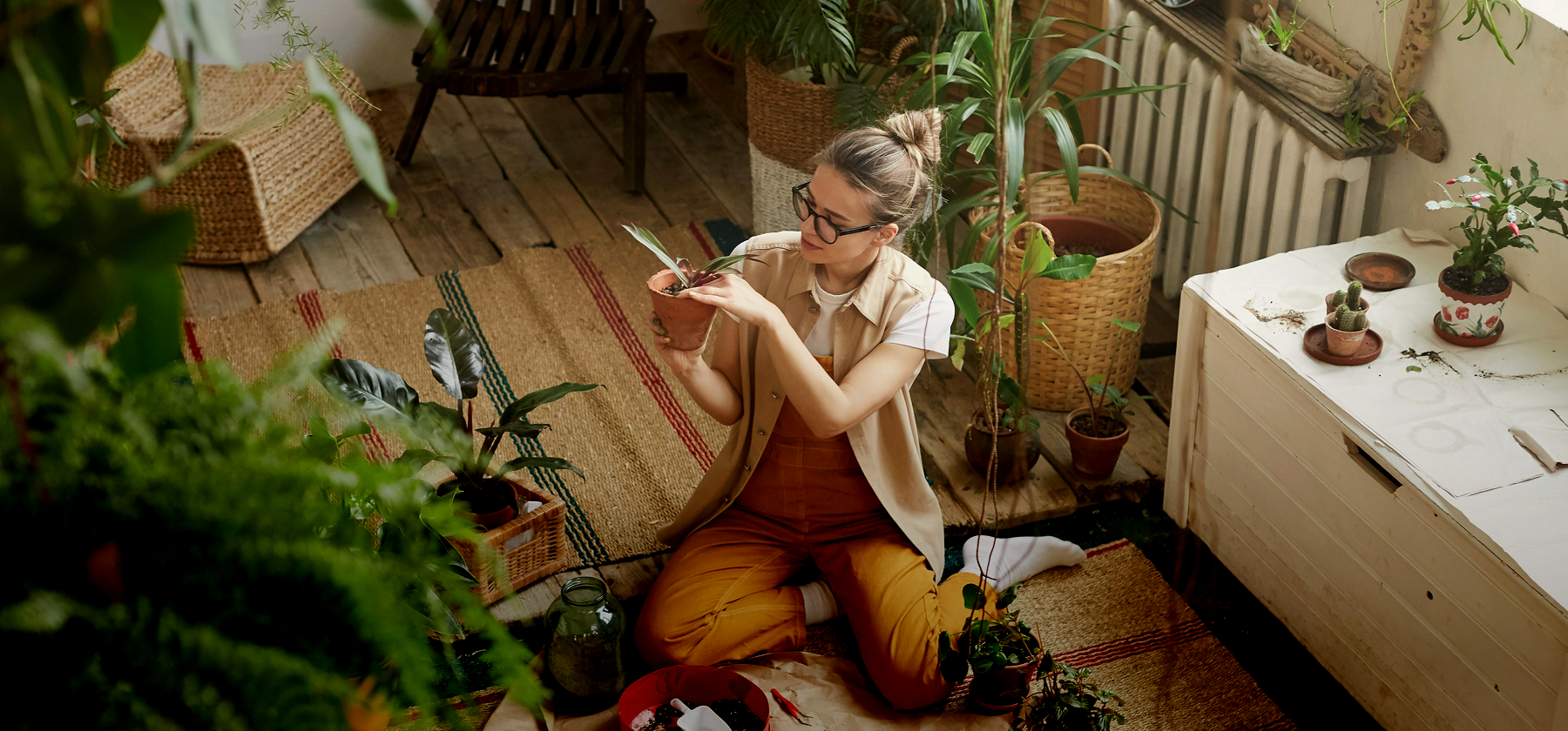 A woman wearing glasses sitting cross-legged on the floor of a plant-filled home, carefully tending to a potted plant surrounded by lush indoor greenery in Germany.