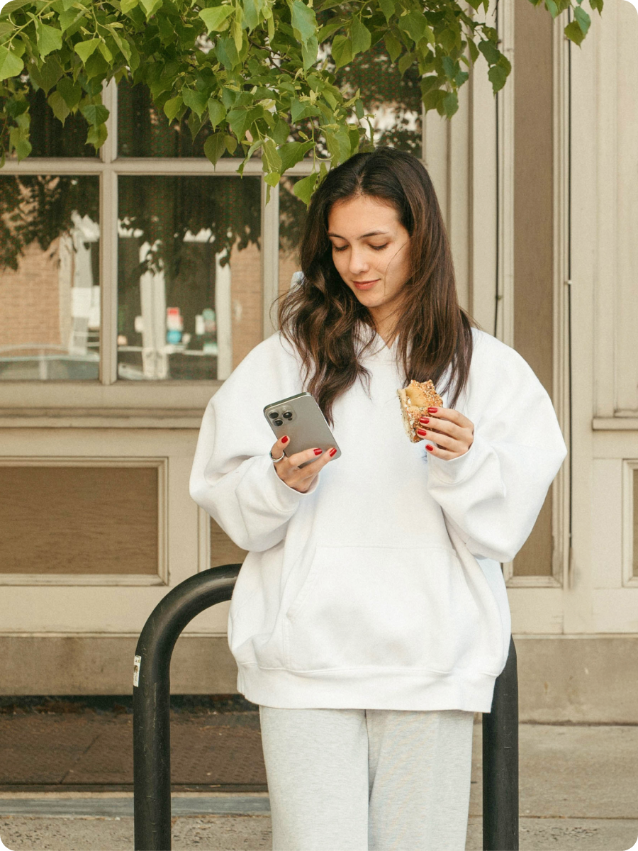 A girl buying Feather insurance on her phone. She stands outside, in front of an apartment building, she's eating a pastry.