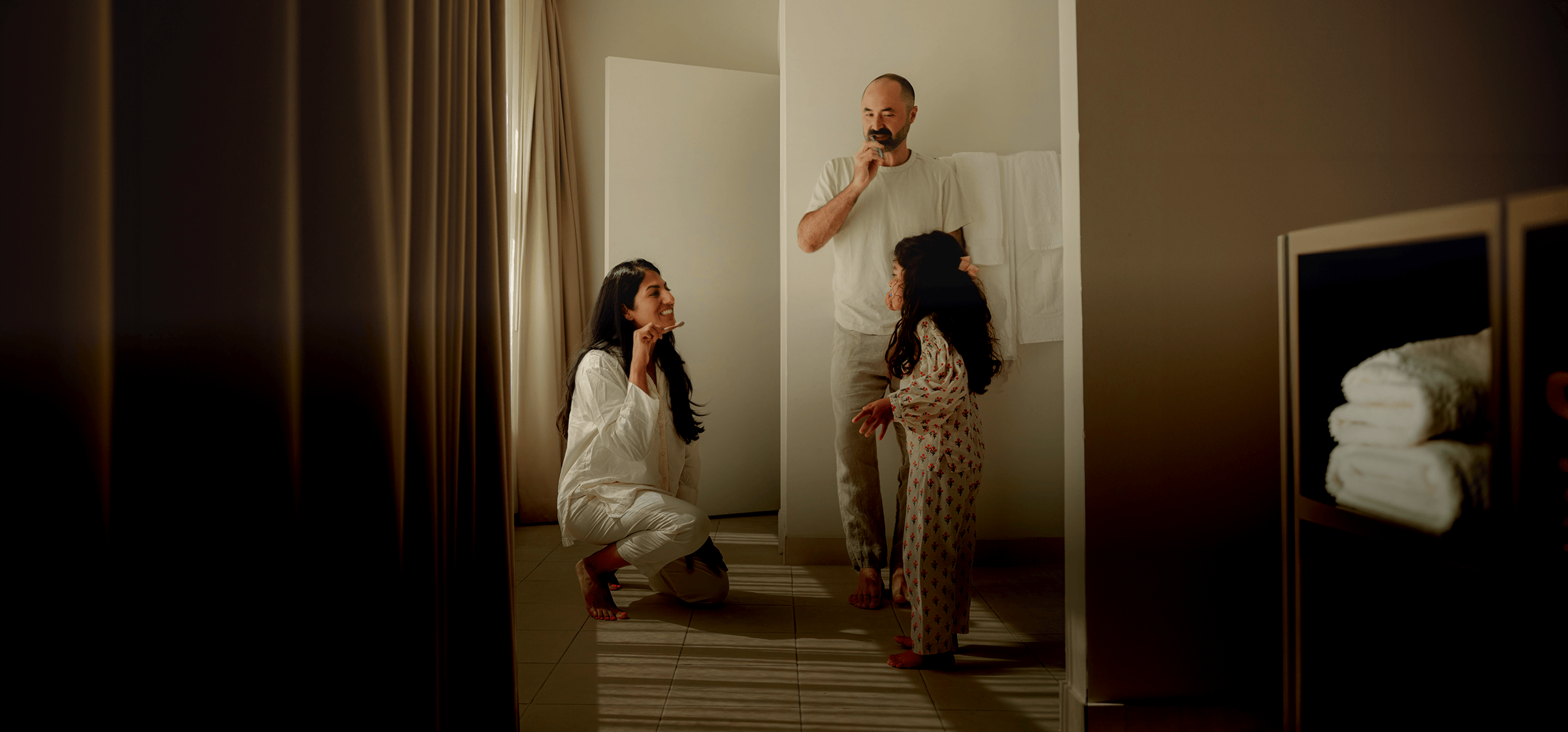 A family brushing their teeth together in a warmly lit bathroom — a mother crouching down to her child's level while a father stands nearby.