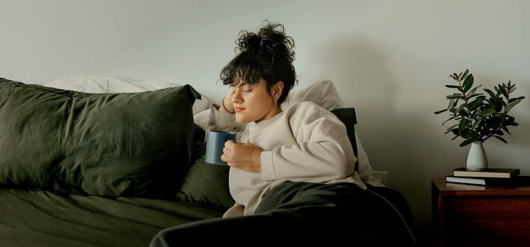  A woman with curly hair sitting in her bed, holding a mug and resting in a calm bedroom in Germany.