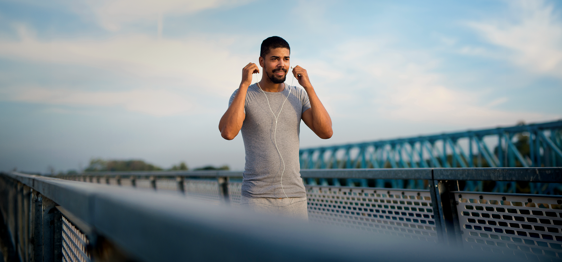 A man in a grey t-shirt putting in earphones while standing on a bridge outdoors on a sunny day in Portugal.
