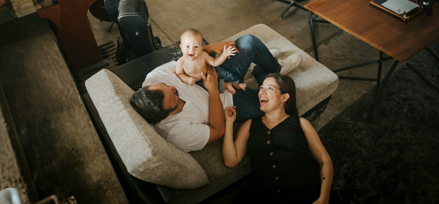 A happy young family with a laughing baby lying on a parent's chest on a sofa, viewed from above, in Portugal.