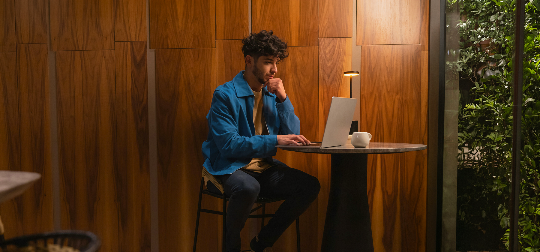 A young man in a blue jacket sitting thoughtfully at a high table with a laptop and coffee cup in a warmly lit, wood-panelled café in Hungary.