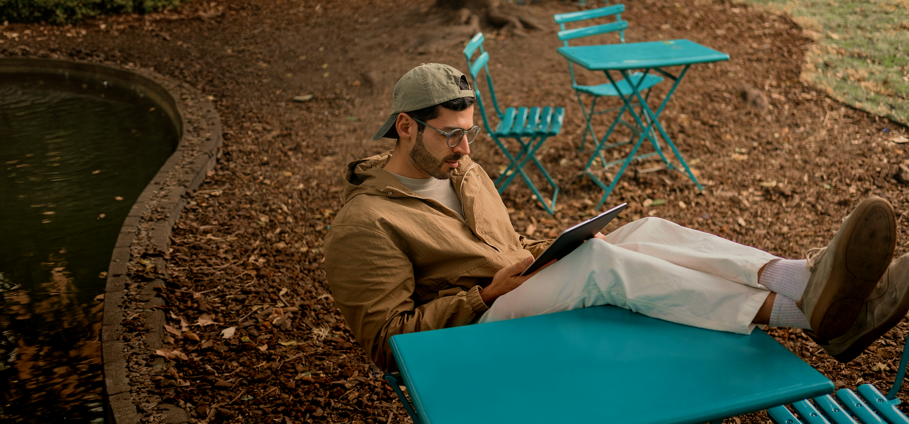 A man wearing glasses and a cap relaxing on a teal outdoor chair in an autumn park, reading on a tablet beside a small pond in Lithuania.