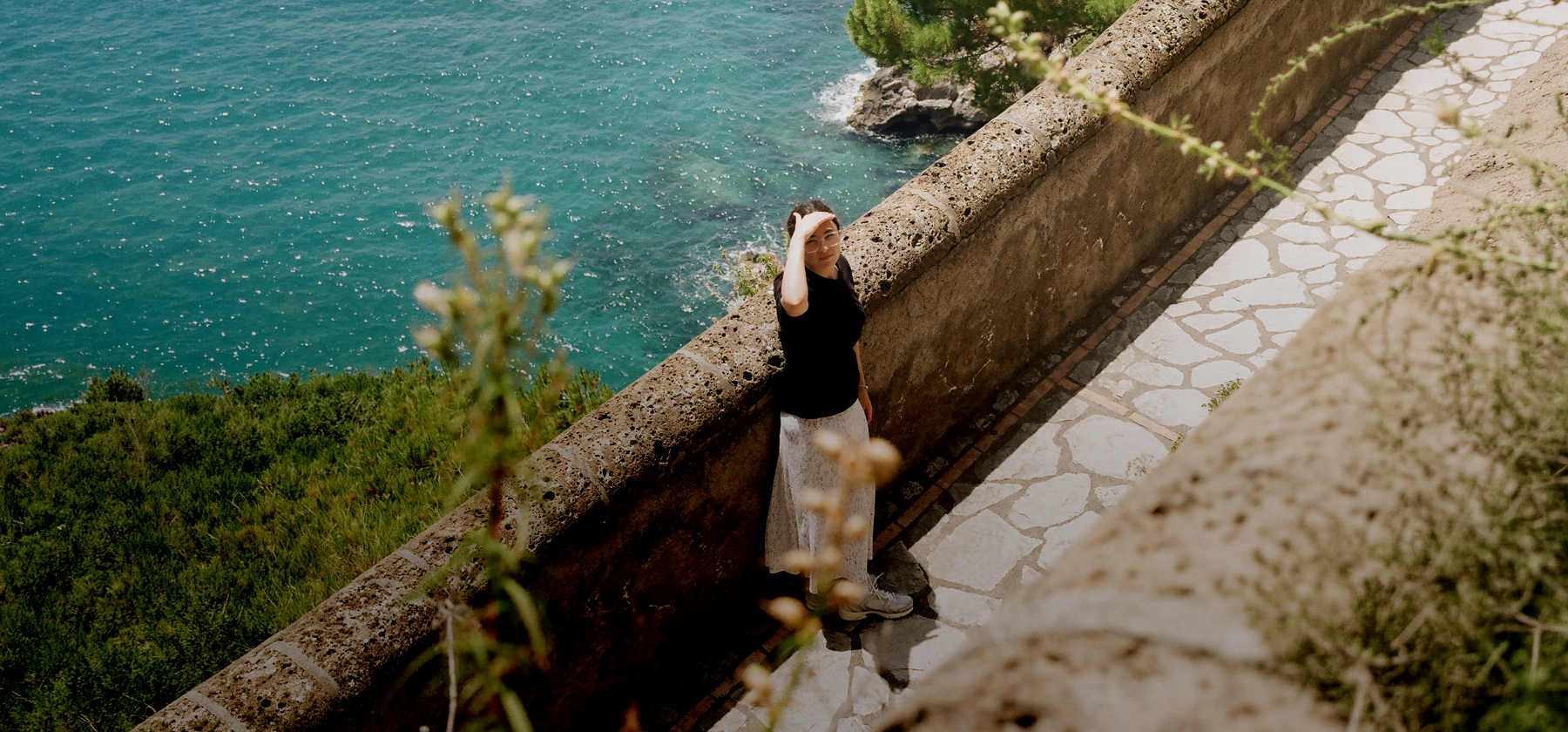 A woman in a black top and white skirt standing on a stone coastal path, looking up at the camera with turquoise Mediterranean waters visible below in Malta.