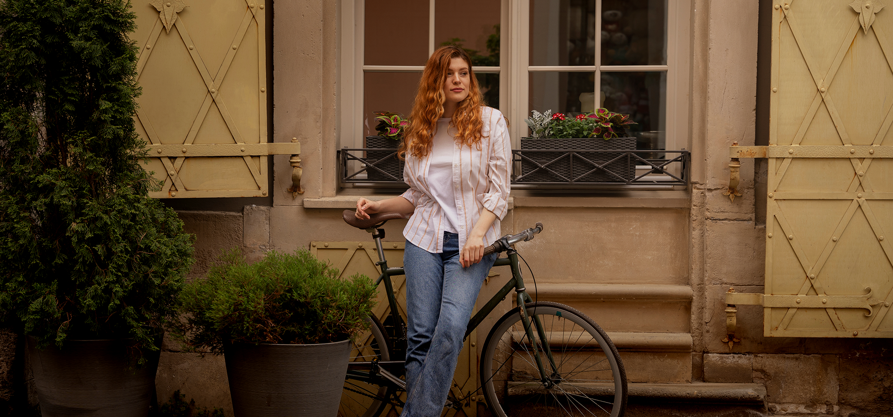 A young woman with long red hair standing with a bicycle in front of a charming European building with flower boxes and shuttered windows in the Netherlands.