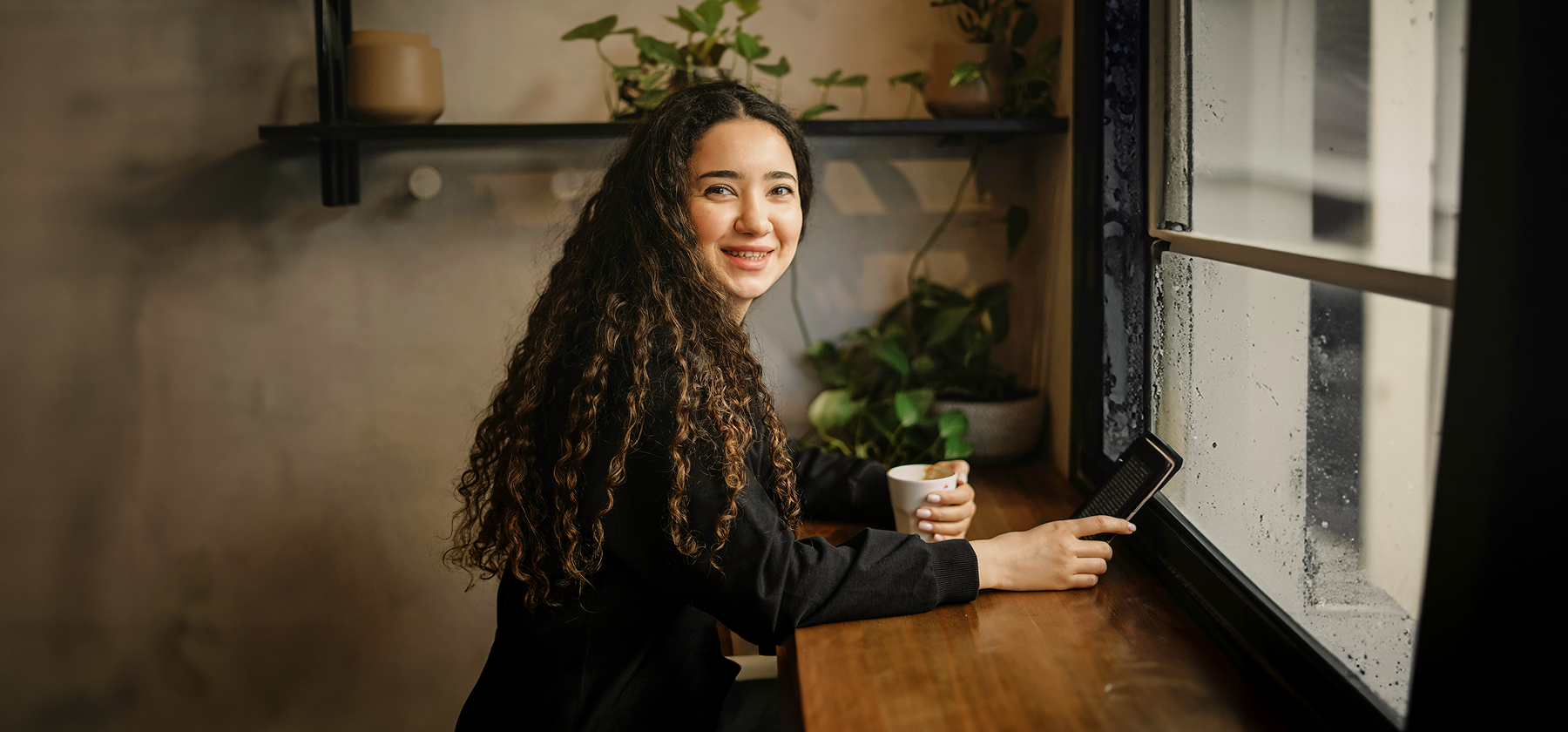 A young woman with long curly hair smiling while holding a coffee cup and using a tablet at a café window in France.