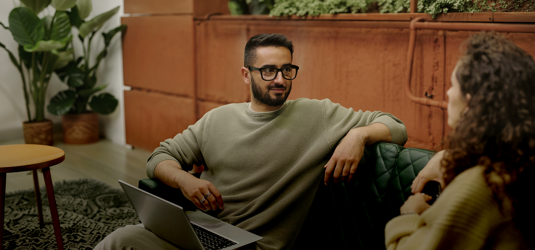 A bearded man wearing glasses sitting on a sofa with a laptop, engaged in conversation with someone across from him in Portugal.