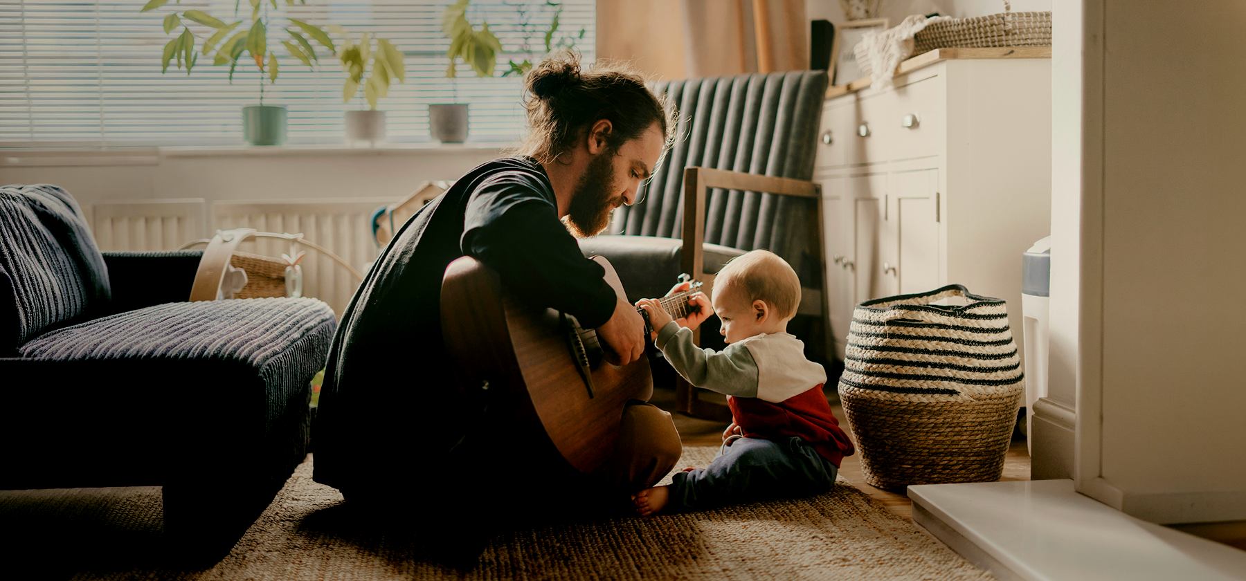 A bearded father sitting on the floor playing guitar for his curious baby in a cosy sunlit living room in France