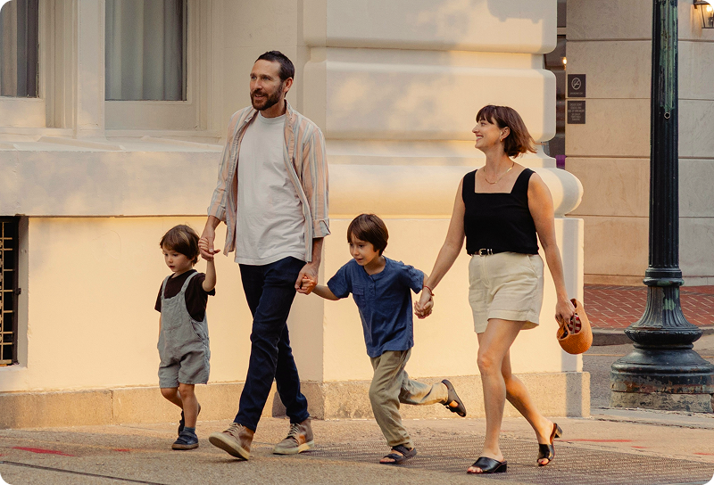 A family of four holding hands and walking together along a sunlit city street, with two young children between their parents in Germany.