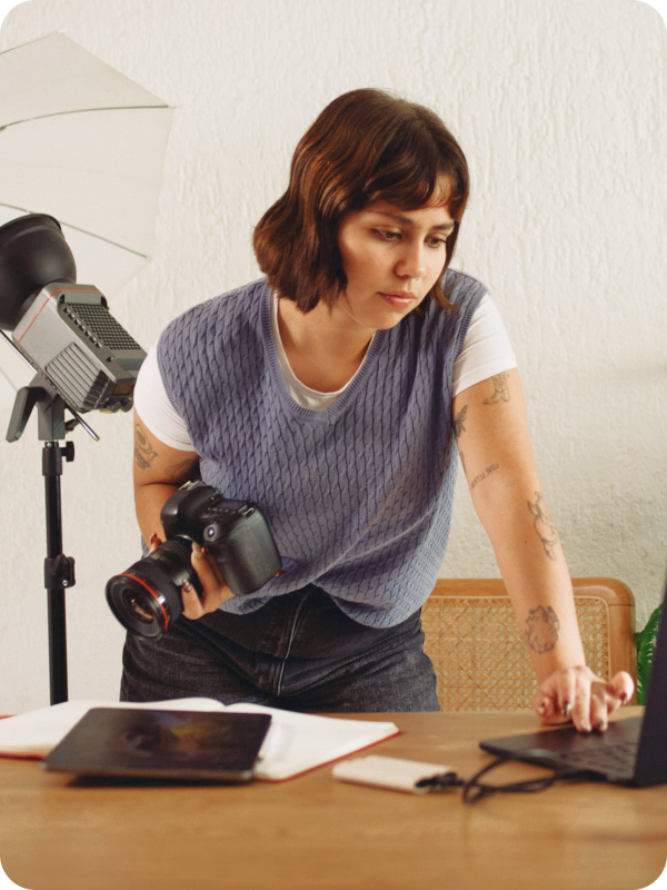 Photographer standing in front of her desk, holding a camera and checking her work on a laptop. Photoshoot equipment is seen in the background of the image.