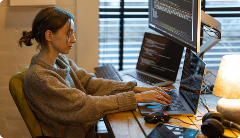 An IT freelancer working at her desk in front of 2 laptops and a screen showing lines of code.