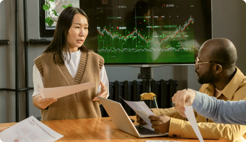 A consultant talking to her client during a business meeting. The tv screen behind her shows graphs. 