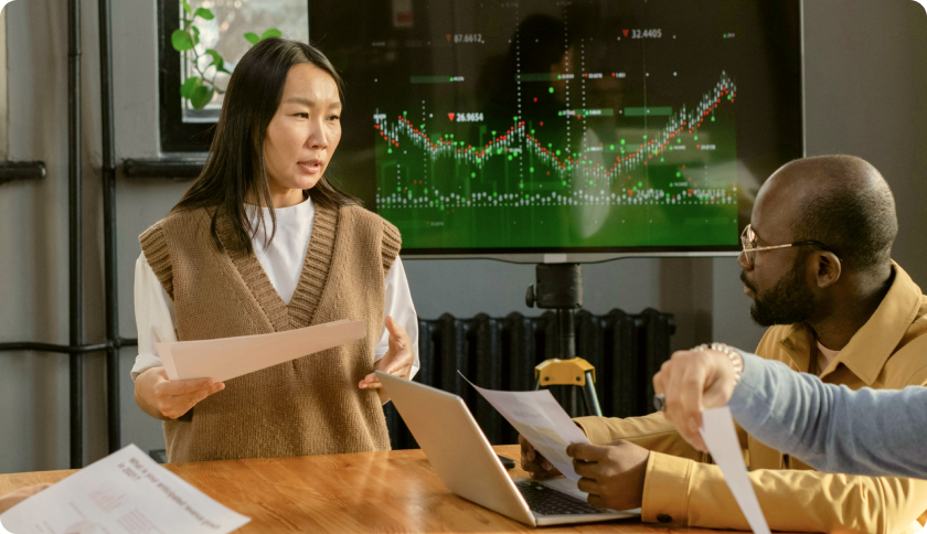 A consultant talking to her client during a business meeting. The tv screen behind her shows graphs.