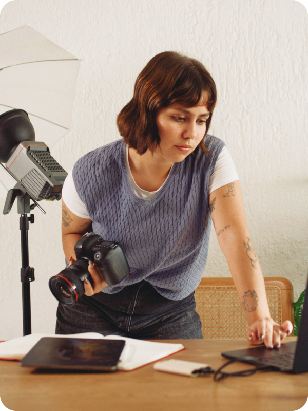 Photographer standing in front of her desk, holding a camera and checking her work on a laptop. Photoshoot equipment is seen in the background of the image.