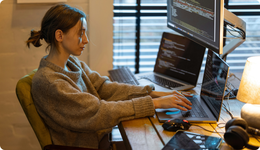 An IT freelancer working at her desk in front of 2 laptops and a screen showing lines of code.
