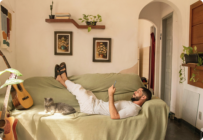 Person laying on a couch at their cosy home, looking at their phone with a cat laying by their side.