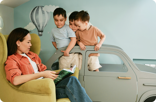 A woman sitting on an armchair reading a story to her 3 toddlers. 