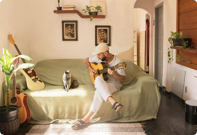 Un homme assis sur un canapé dans un appartement cosy, jouant de la guitare. Un chat gris est assis à côté de lui.