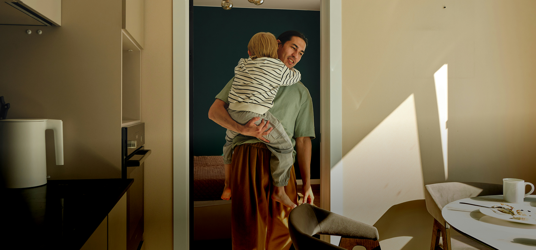 A father holding a young child in his arms in a sunlit modern apartment kitchen in France.