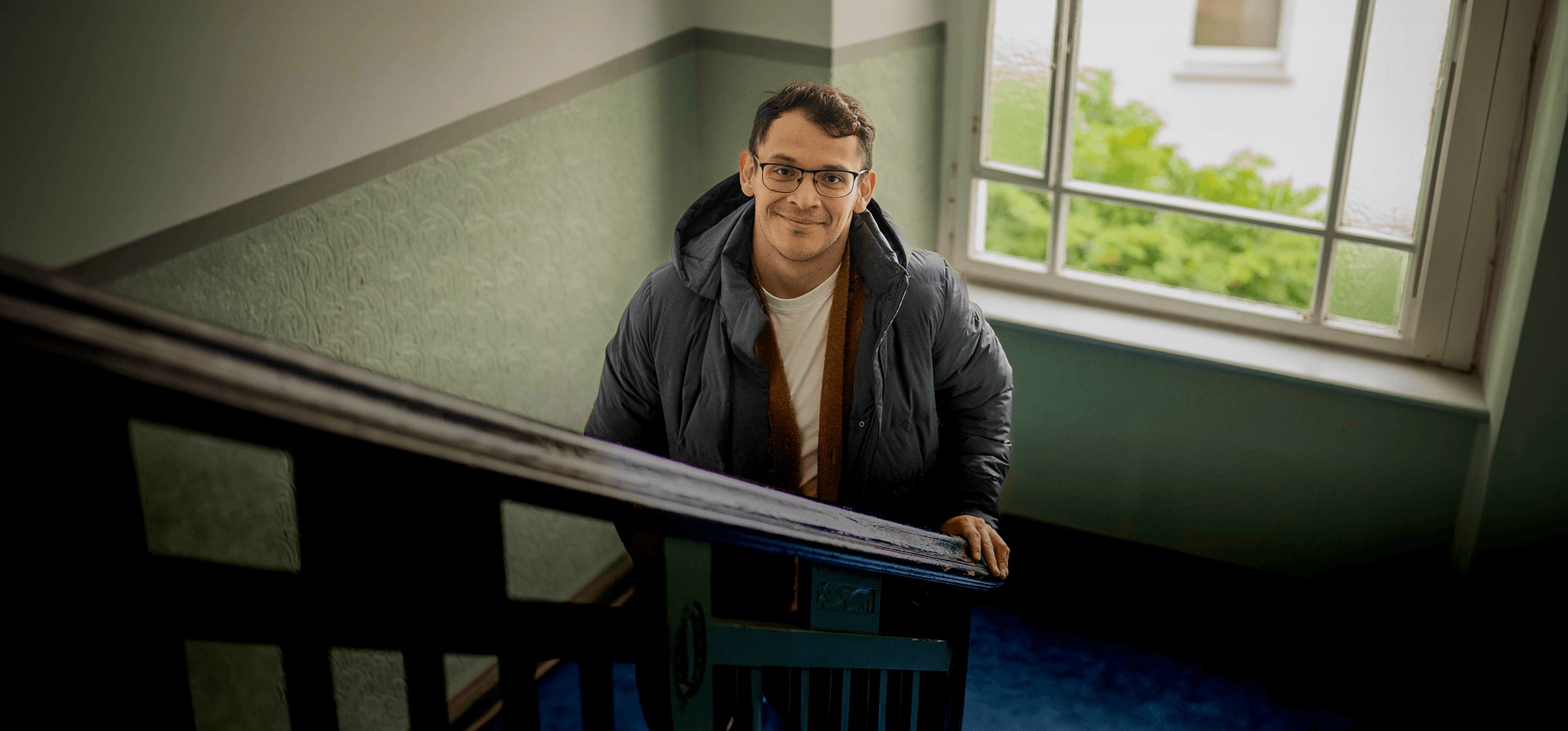 A man wearing glasses and a dark puffer jacket looking up at the camera while climbing a staircase in an apartment building in France.