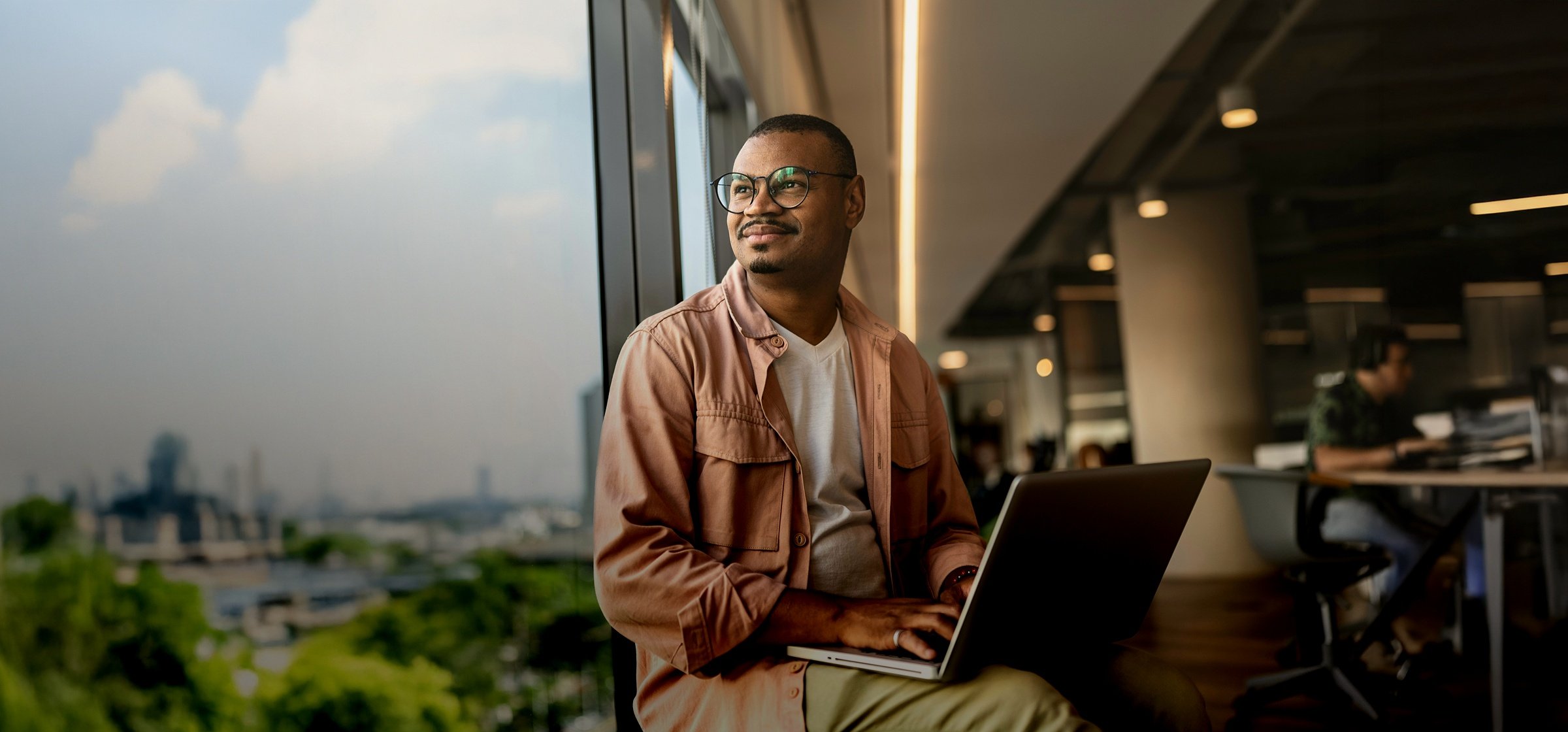 A man with glasses sitting with a laptop by a large office window overlooking a city skyline in Spain